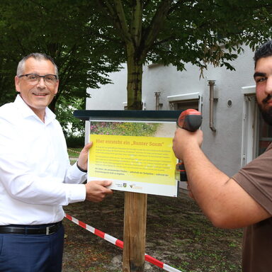 Vizelandrat Andreas Siebert (links) und Hausmeister Yasar Uzal befestigen ein Hinweisschild an der neuen Blühfläche.